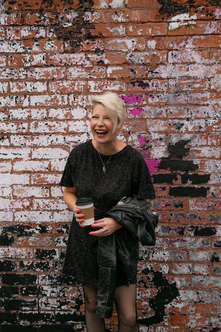 Zoe laughing with a takeaway coffee in hand and a leather jacket over her arm, against a painted brick wall