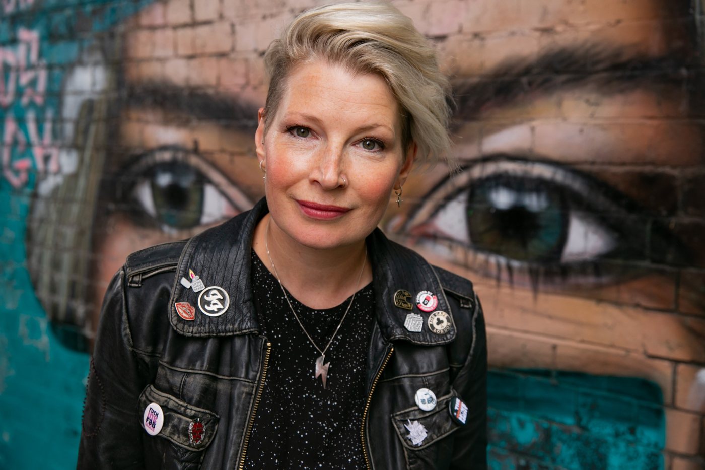 Close portrait of Zoe looking straight at the camera, in a badge-covered leather jacket, in front of a mural of giant painted eyes