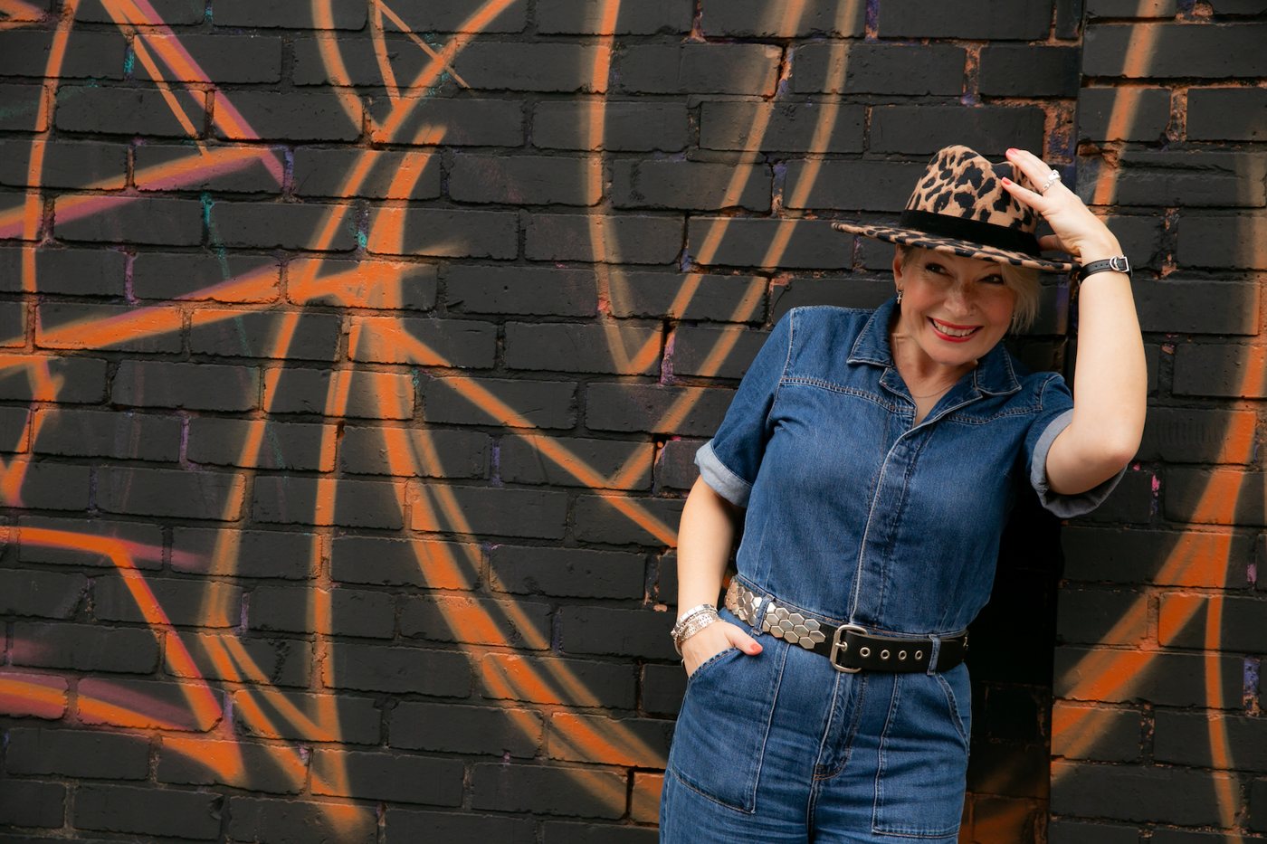 Zoe tipping a leopard print hat and grinning, in a denim jumpsuit against a graffiti wall