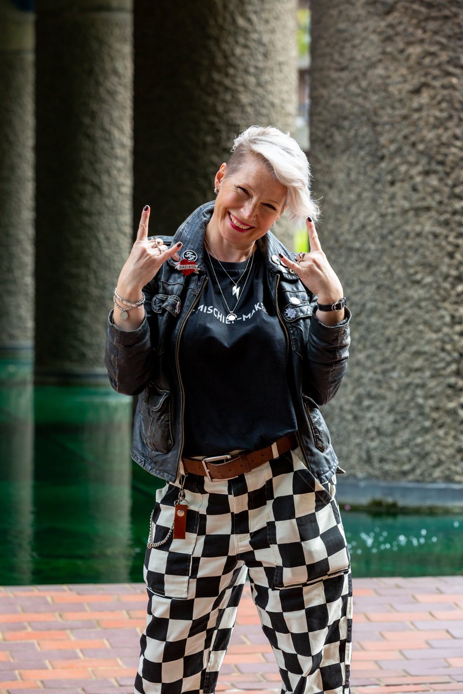 Zoe grinning and throwing rock horns with both hands, in a badge-covered leather jacket and Mischief-Maker tee